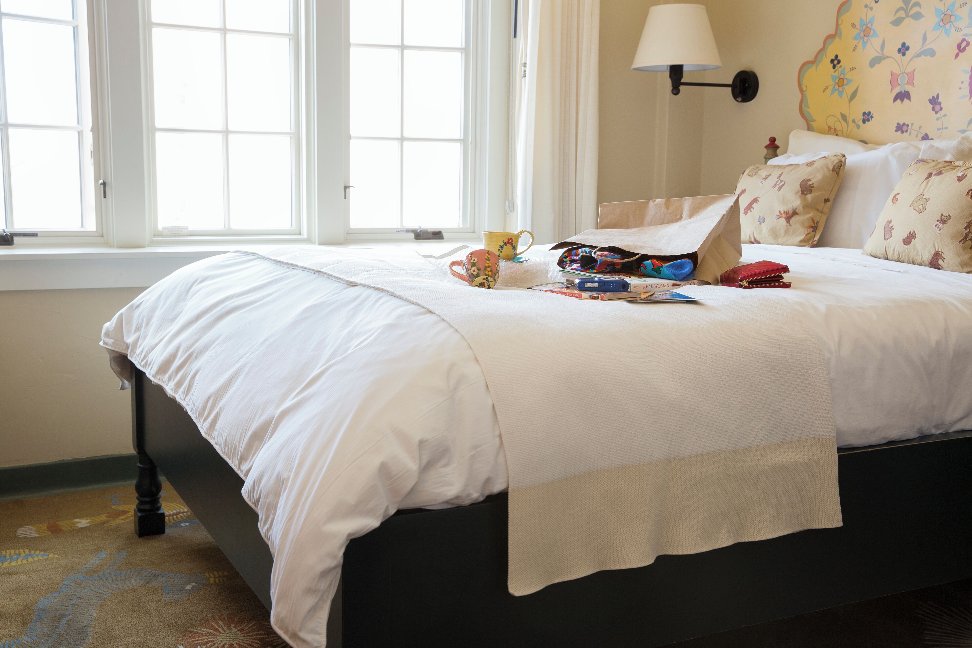 A neatly made bed with a white comforter, a lamp on the wall, and a window with curtains.
