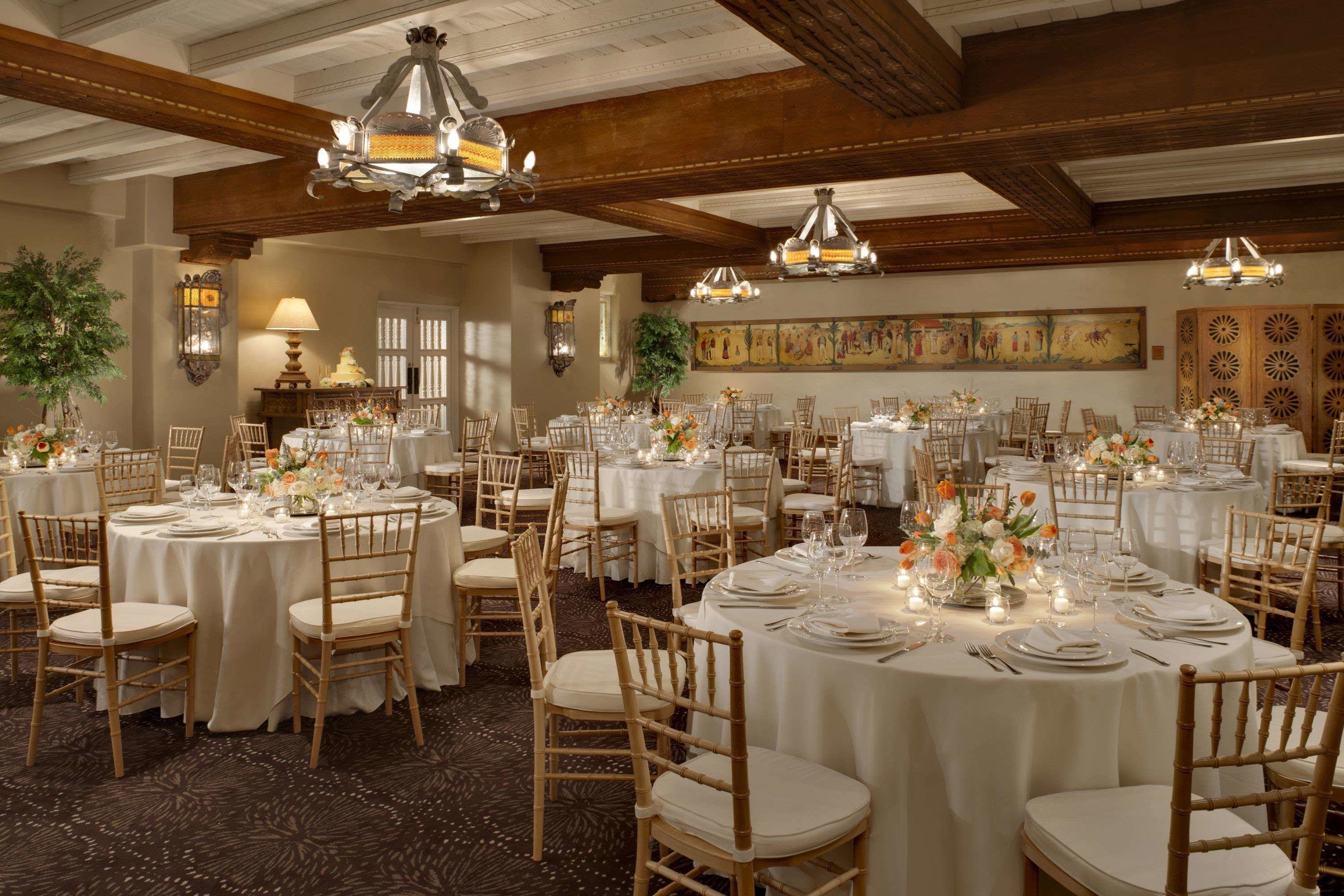 A banquet hall with round tables set for dining, wooden chairs, and chandeliers.