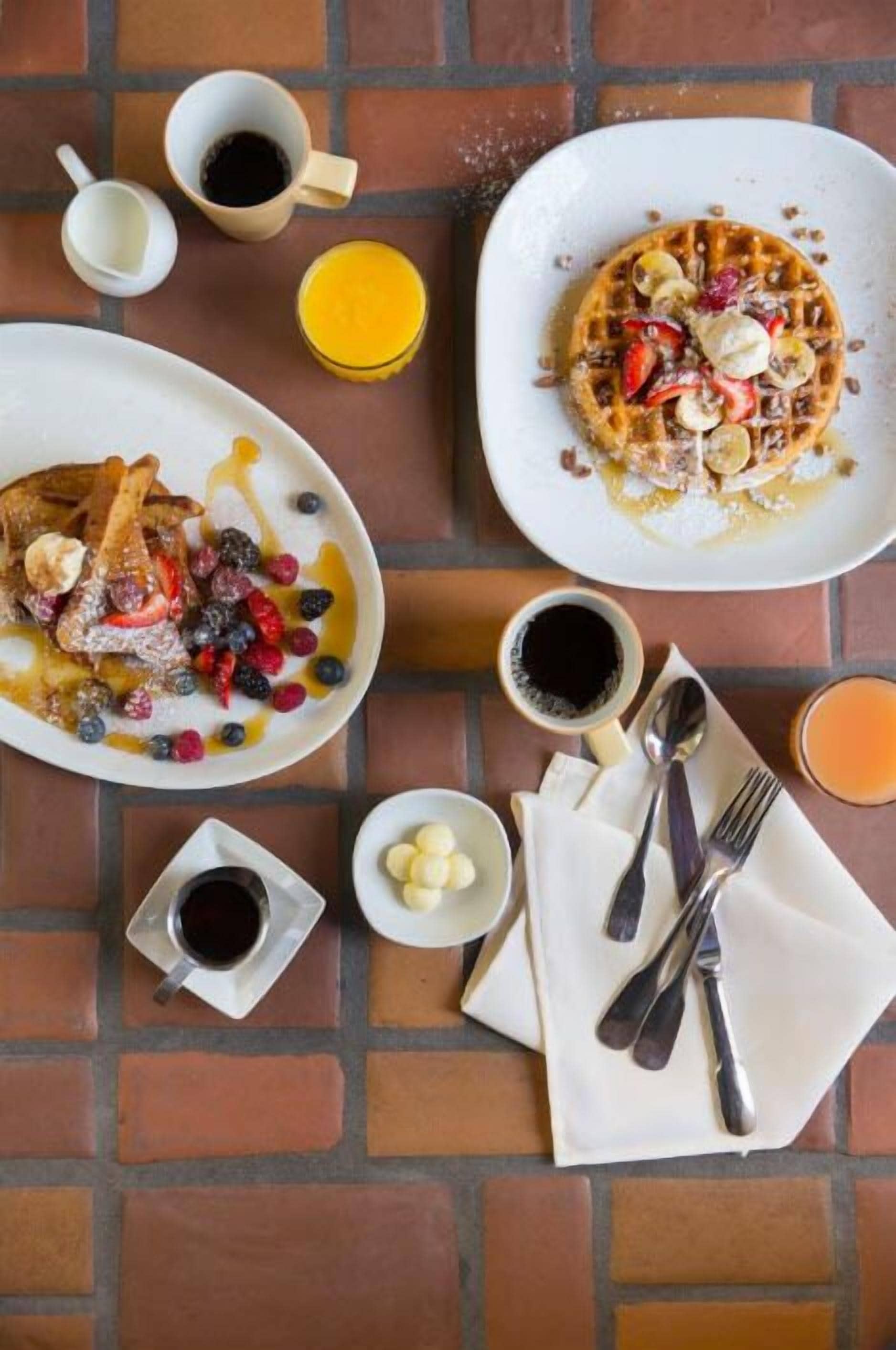 A breakfast setting with waffles, pancakes, and berries on plates, accompanied by cups of coffee and syrup.