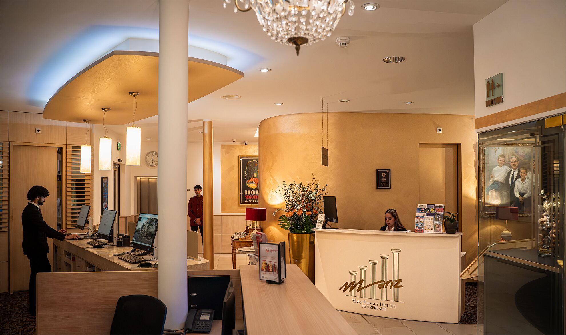 Ein Hotel-Empfangsbereich mit einer gebogenen Rezeption, einer Kristallleuchtre und einer Wanduhr.