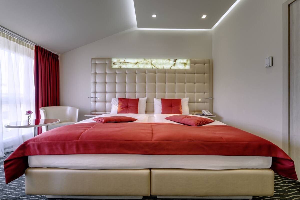 Habitación de hotel moderna con una cama grande, una mesita de noche, una silla y un ventanal con cortinas.