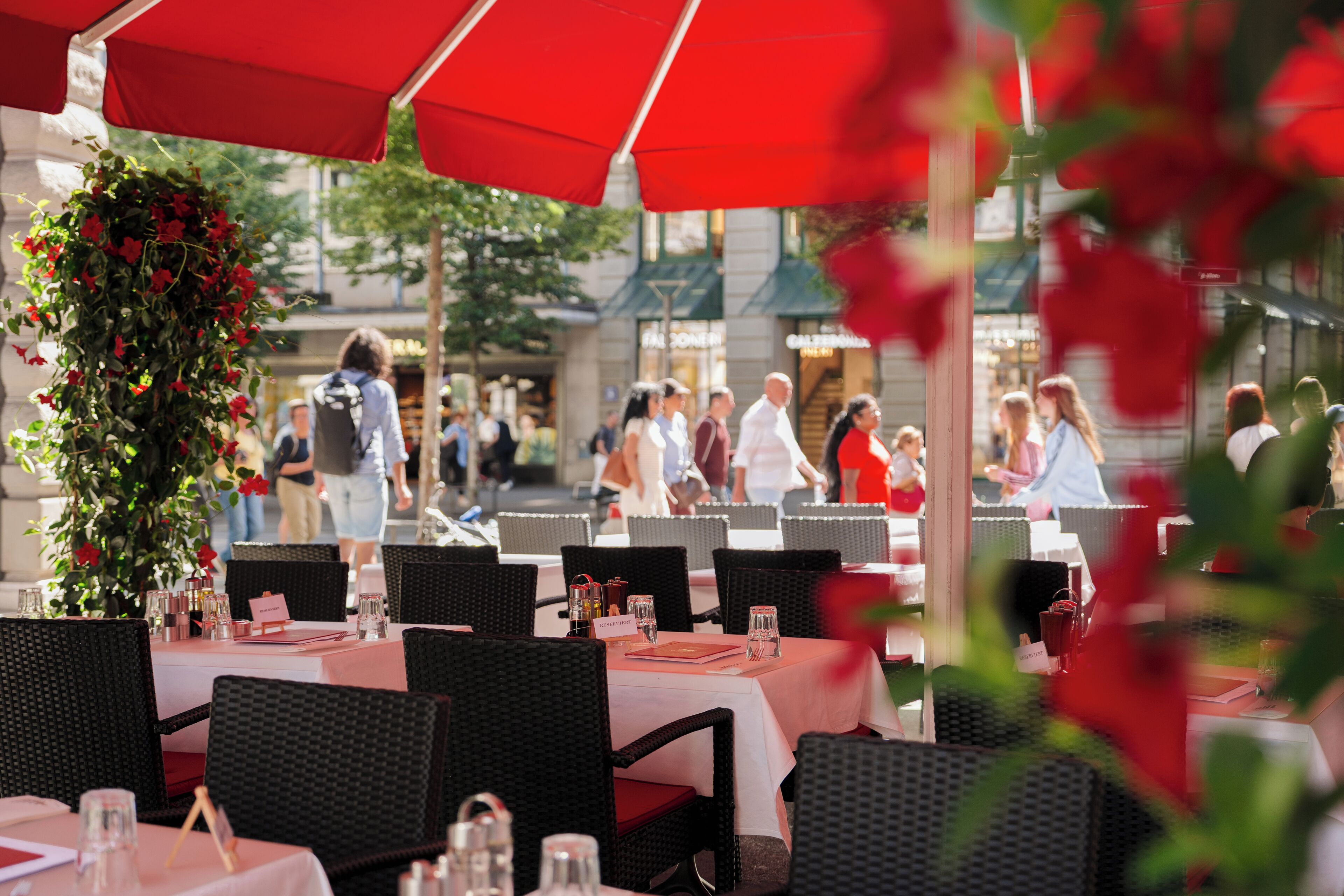 An outdoor dining area with wicker chairs and tables, red umbrellas, and a decorative floral arrangement.