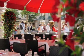 An outdoor dining area with wicker chairs and tables, red umbrellas, and a decorative floral arrangement. - St Gotthard Hotel (Zürich)