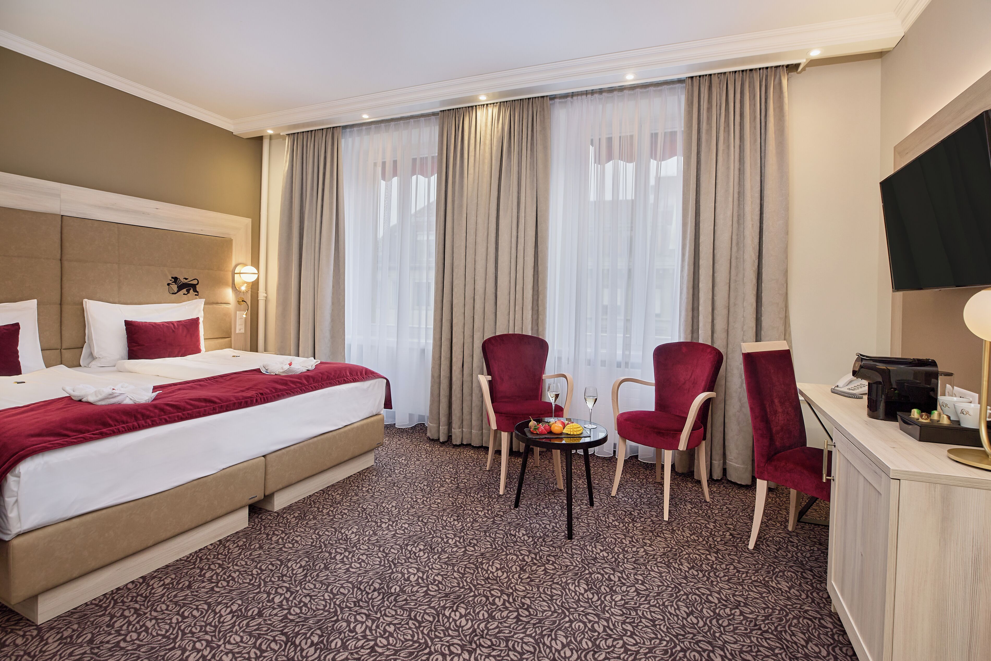 A hotel room with a bed, two red chairs, a small table with a fruit bowl, a coffee machine, and a TV on the wall.