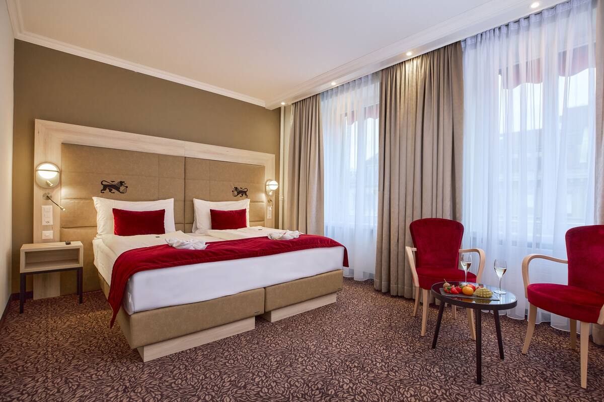 Habitación de hotel con una cama grande, dos sillones rojos, una mesita con fruta y un vaso, y vistas de la ciudad a través de cortinas traslúcidas.