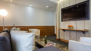 Une chambre d’hôtel moderne dotée d’un téléviseur à écran plat fixé au mur, d’un canapé, d’une petite table avec une bouteille et des verres, et d’un mur revêtu de panneaux en bois.