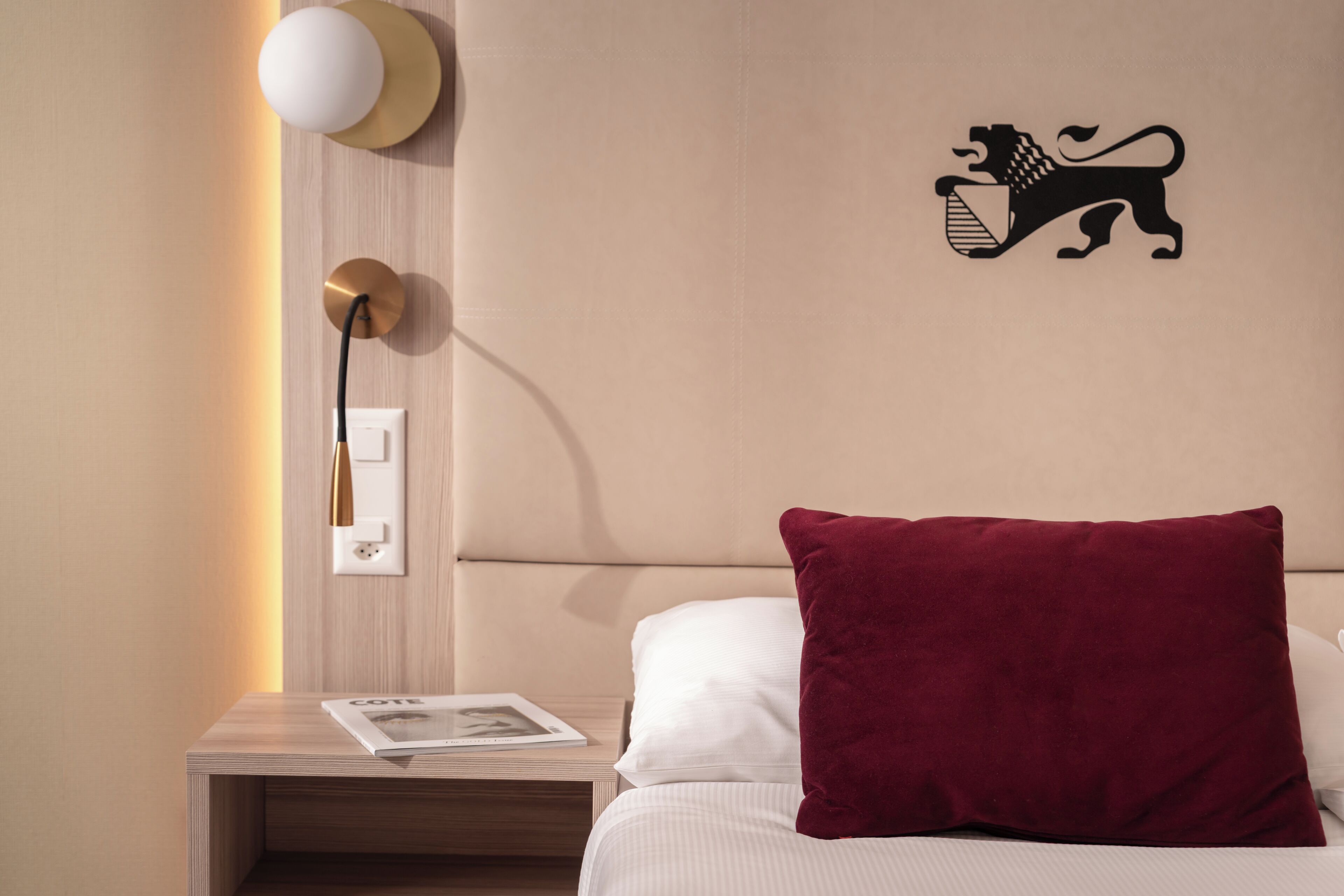 Une chambre d’hôtel comprenant un lit, une table de chevet, une lampe fixée au mur, une prise de courant et un emblème décoratif représentant un lion.