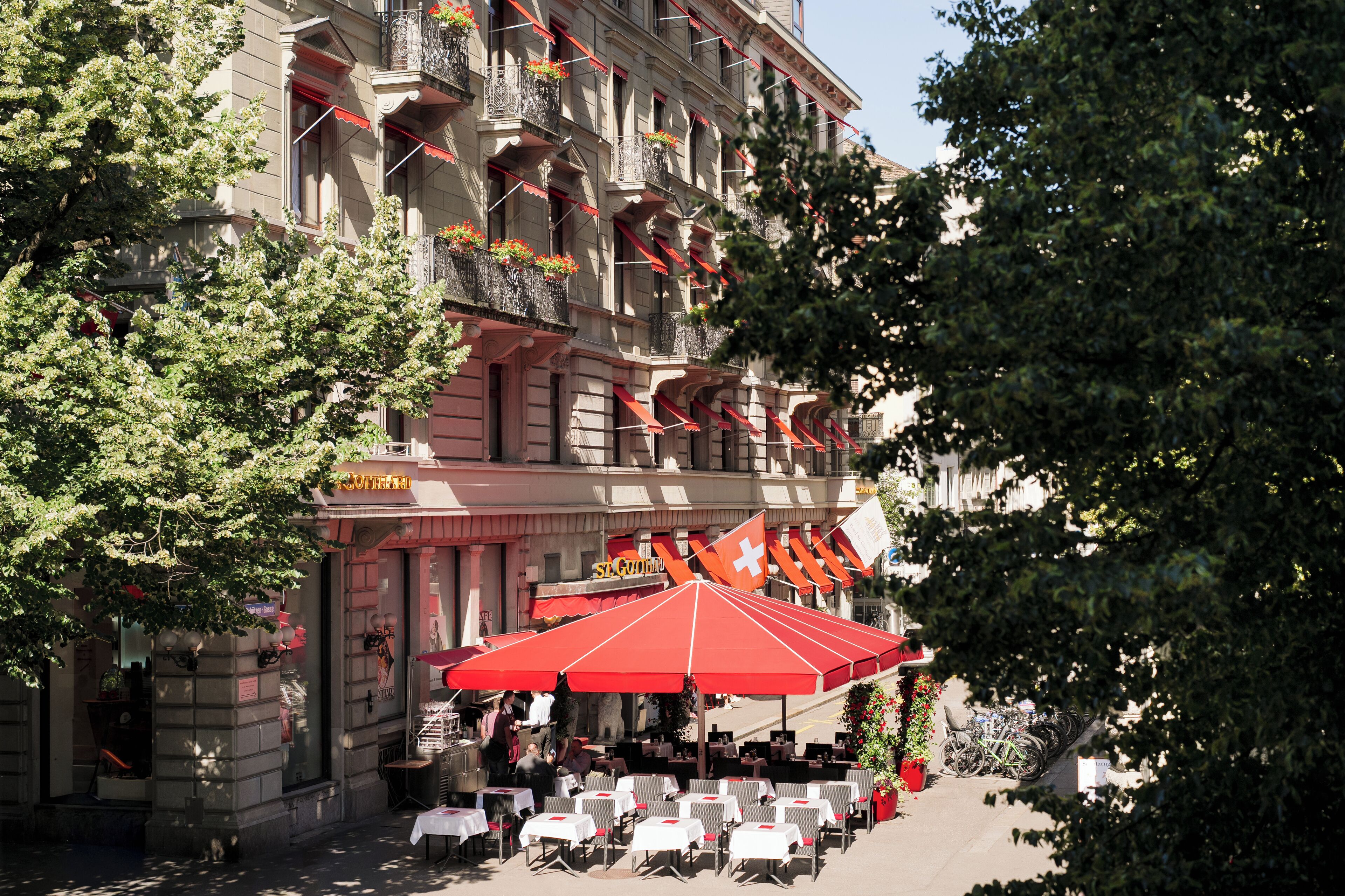 An outdoor café with tables and chairs, a red umbrella, and a building with balconies and flags.