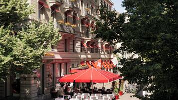 An outdoor café with tables and chairs, a red umbrella, and a building with balconies and flags.