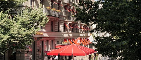 Un café al aire libre con mesas y sillas, un paraguas rojo, y un edificio con balcones y banderas.