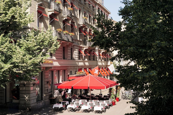 Un café al aire libre con mesas y sillas, un paraguas rojo, y un edificio con balcones y banderas.