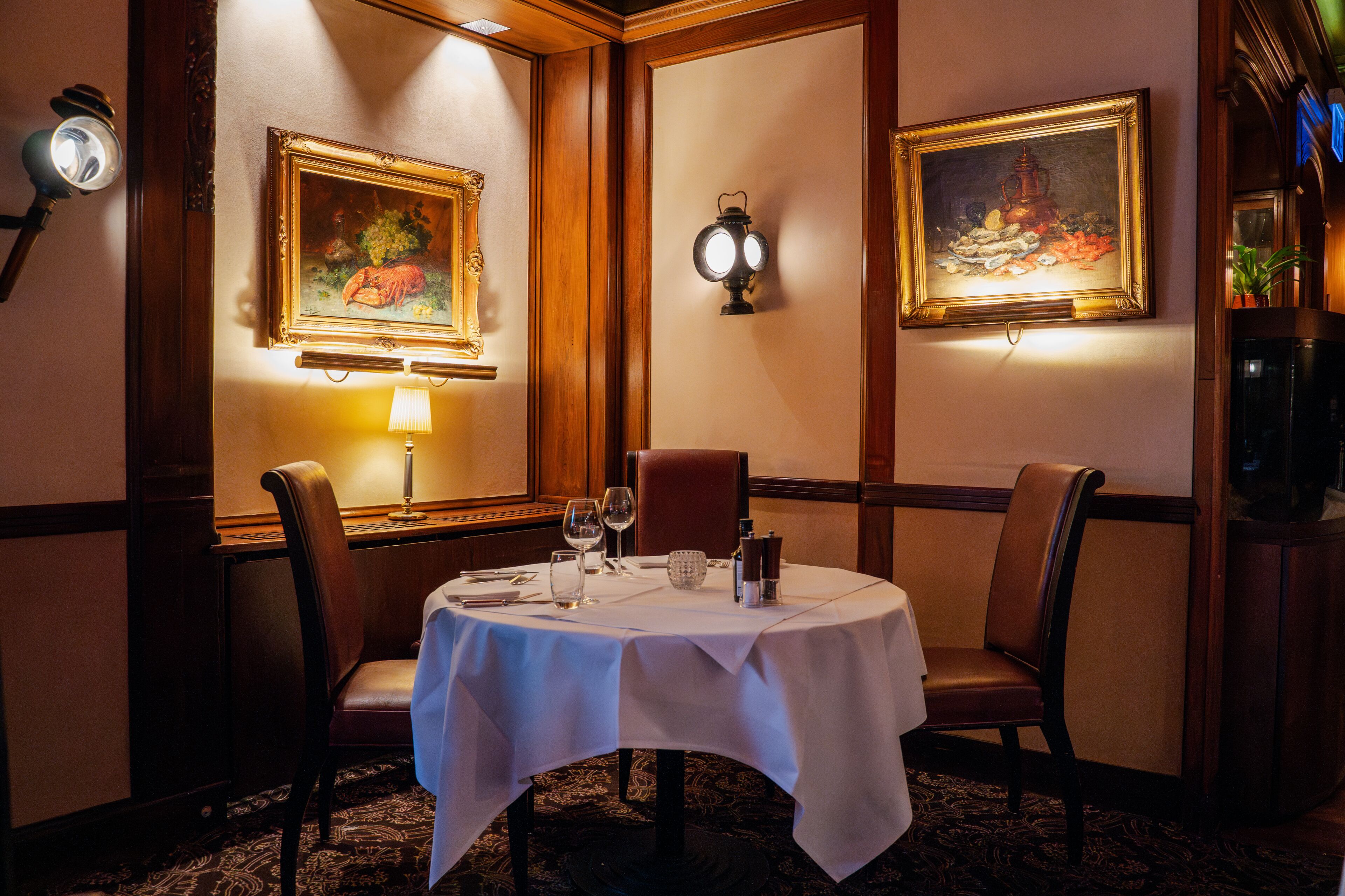A dining area with a table set for two, chairs, a wall-mounted light, and two framed paintings.