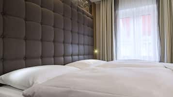 Una habitación de hotel con cabecera capitonada, una cama grande con ropa blanca y una ventana con cortinas de encaje.
