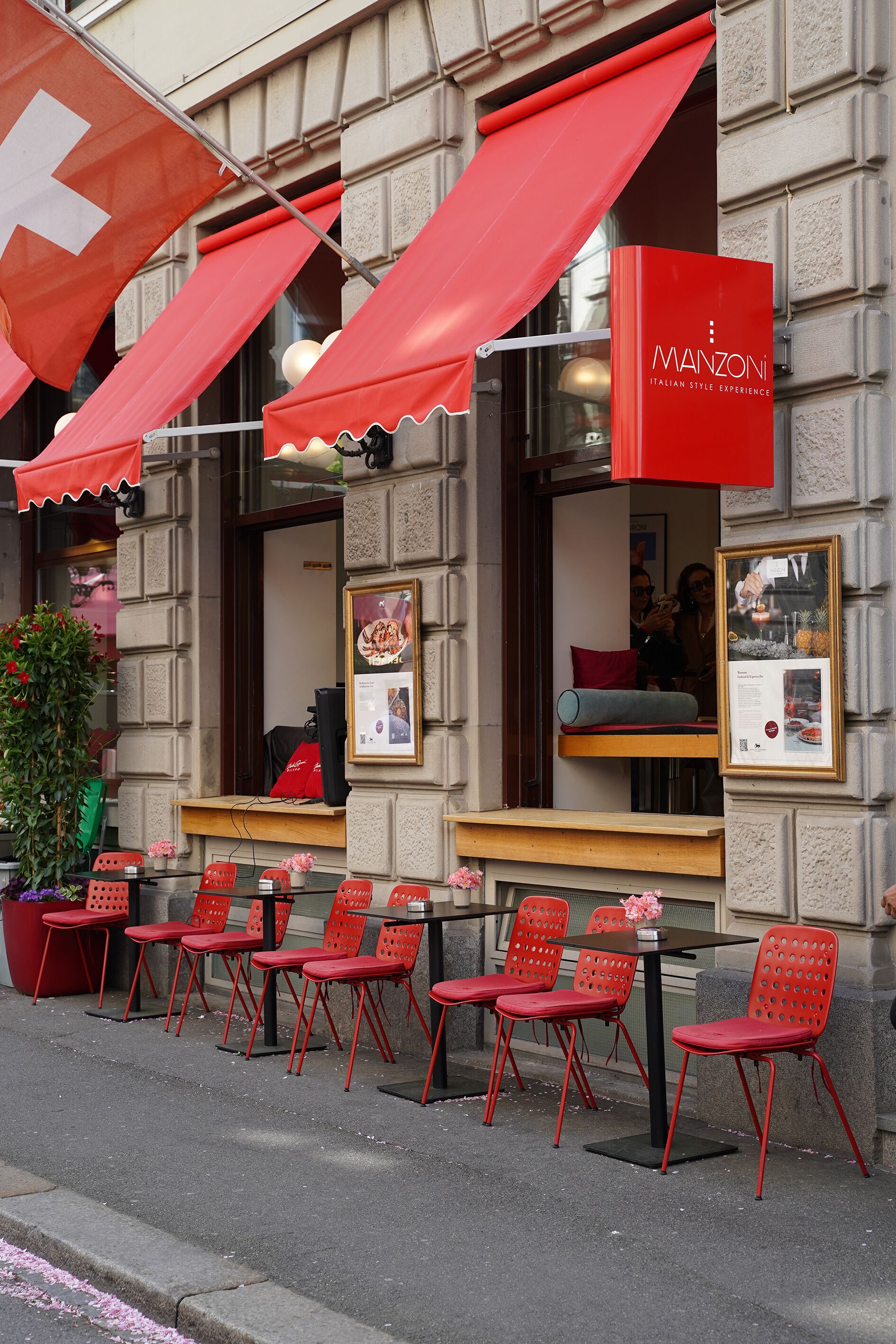 Un área de estar al aire libre con sillas y mesas rojas, una planta en maceta y un toldo rojo con el texto 'MANZONI ITALIAN STYLE EXPERIENCE'.