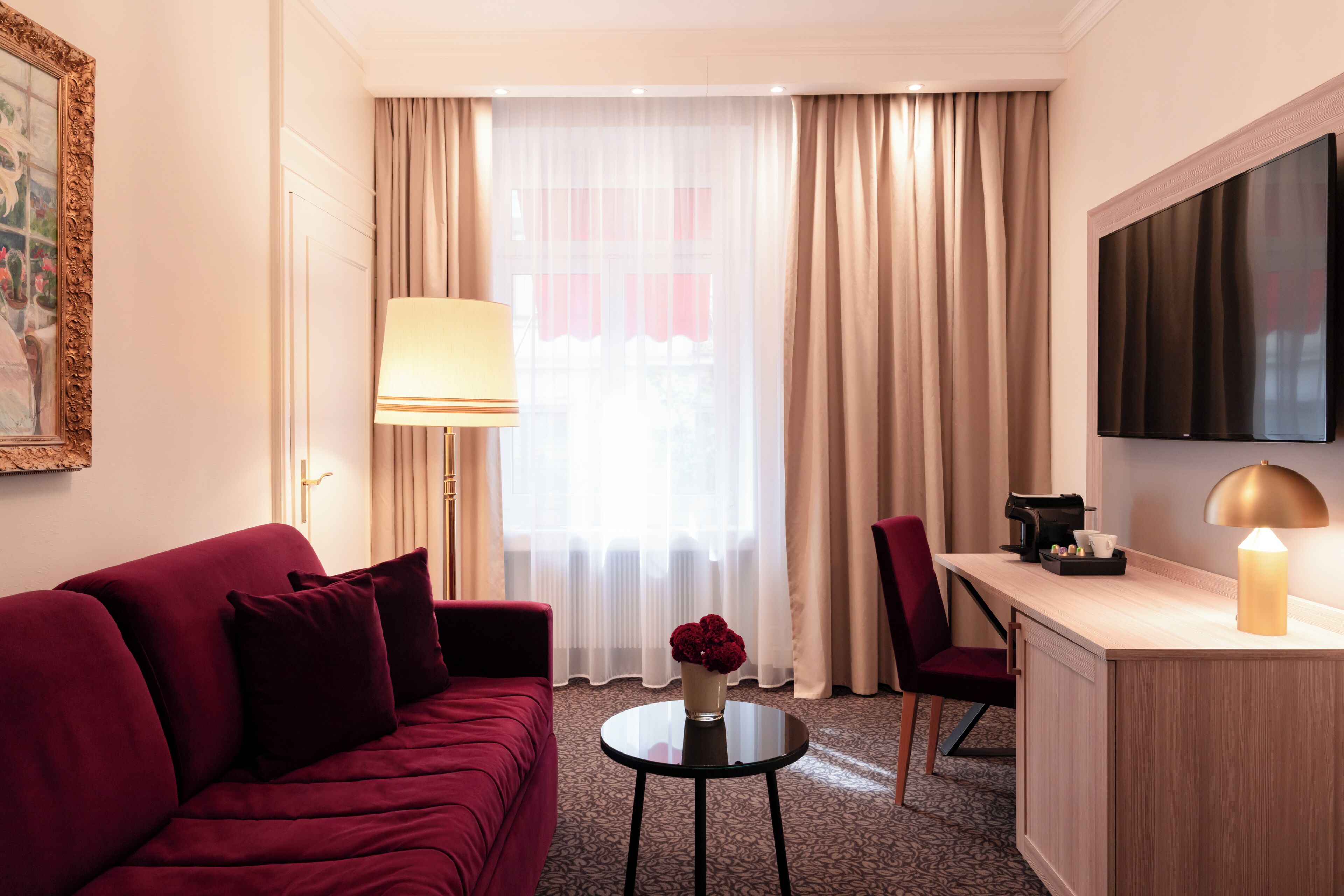 A hotel room with a red sofa, a small round table, a desk with a coffee maker, a TV mounted on the wall, and a window with sheer curtains.