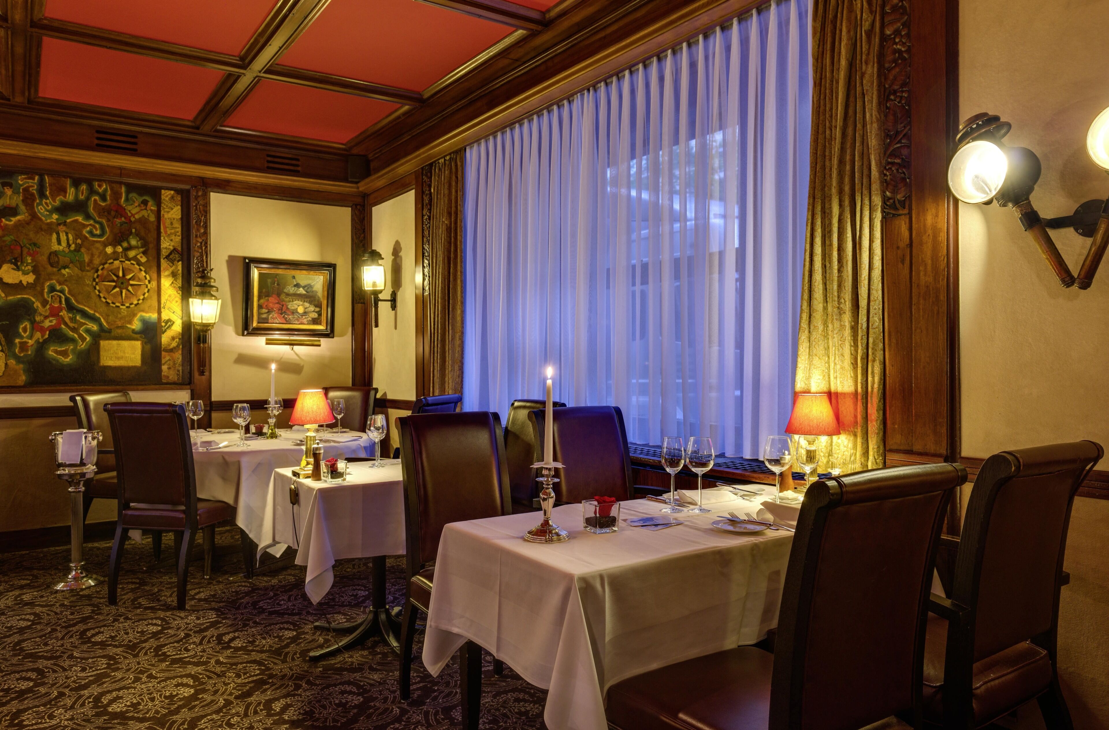 Ein Hotelrestaurant mit Tischen für zwei Personen, eleganten Stühlen, einem großen Fenster mit Vorhängen und Wandleuchten.