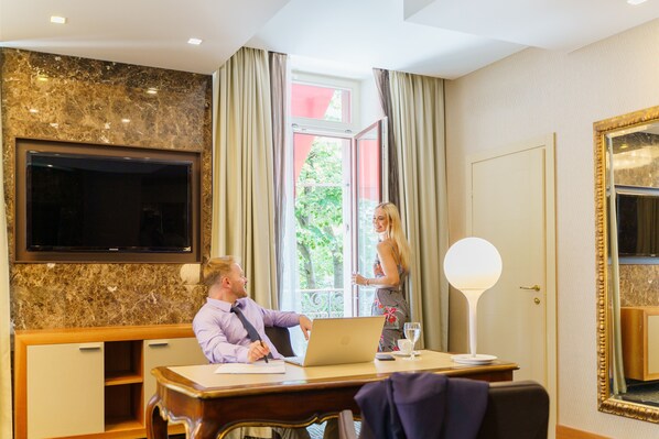 Quarto de hotel com uma televisão de tela plana grande fixada na parede, uma mesa com um laptop, um abajur e uma janela grande com cortinas.