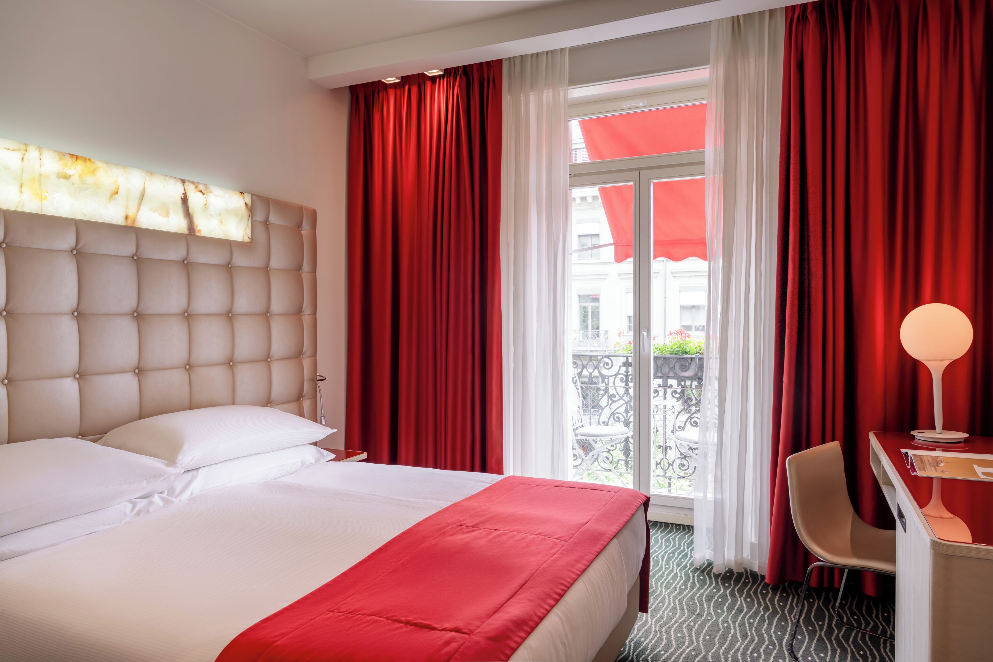 Ein Hotelzimmer mit einem großen Bett, einem Schreibtisch mit Lampe und Blick ins Freie durch ein Fenster mit roten Vorhängen.