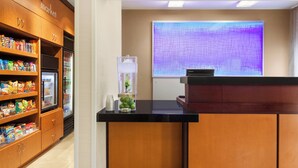 Een hotel receptie met een houten kast vol snacks, een koelkast, een kleine tafel met een plant en een scherm dat een paars abstract patroon projecteert.