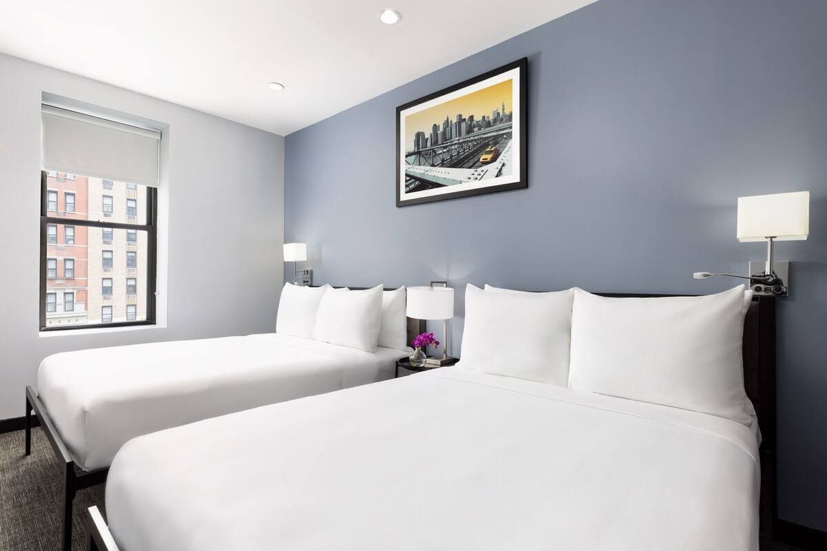 A hotel room with two beds, a cityscape painting, a window with a view of buildings, and a nightstand with a lamp.