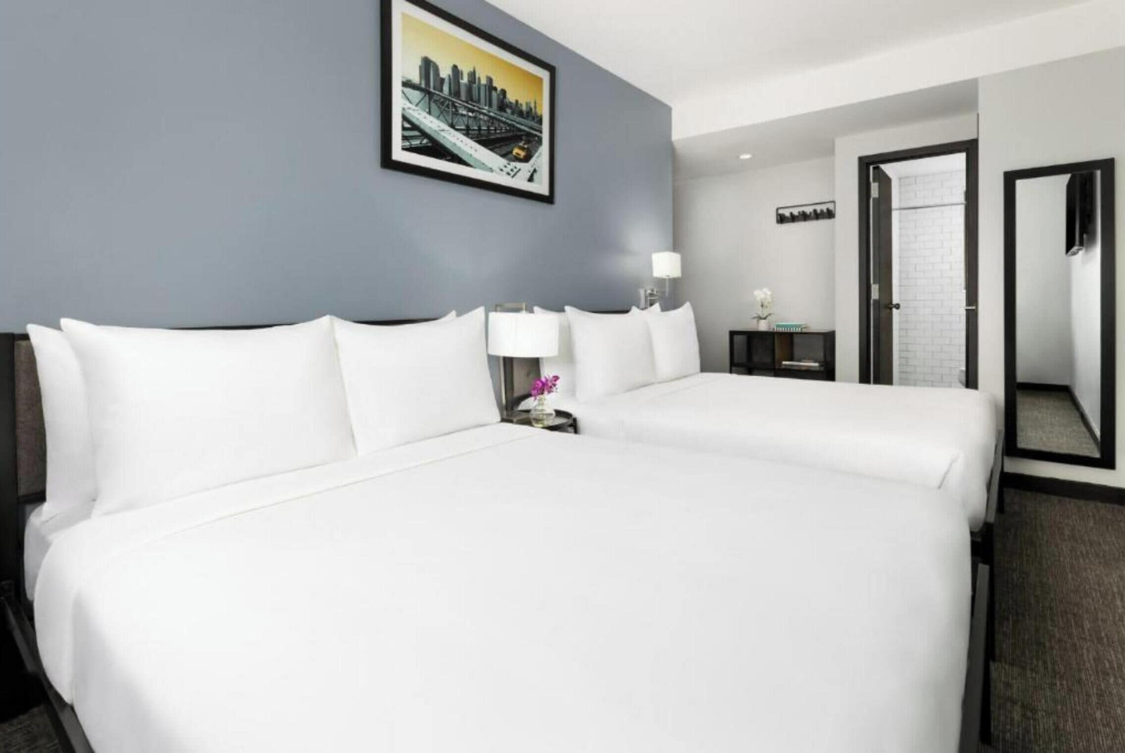 Habitación de hotel con dos camas, un cuadro de un paisaje urbano en la pared, una mesita de noche con una lámpara y una puerta que da a otra habitación.