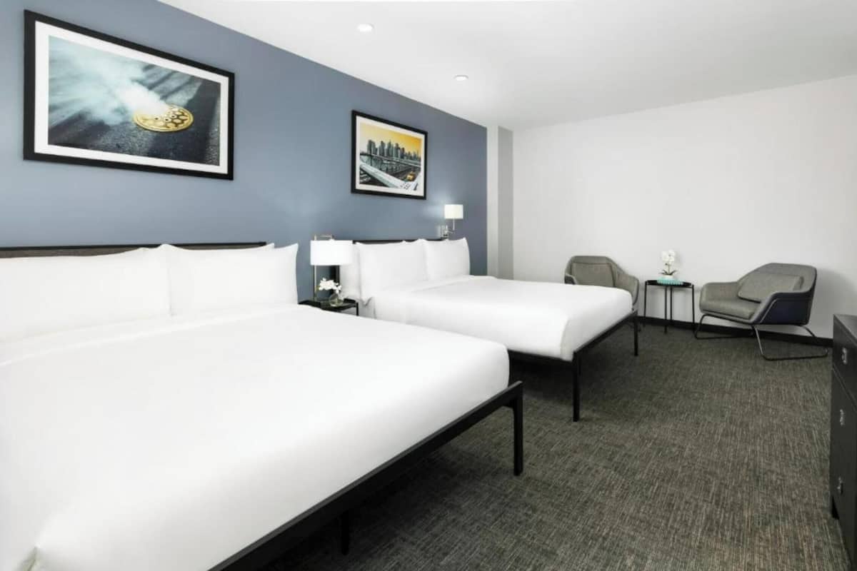 A hotel room with two beds, a gray carpet, a chair, and two framed pictures on the wall.