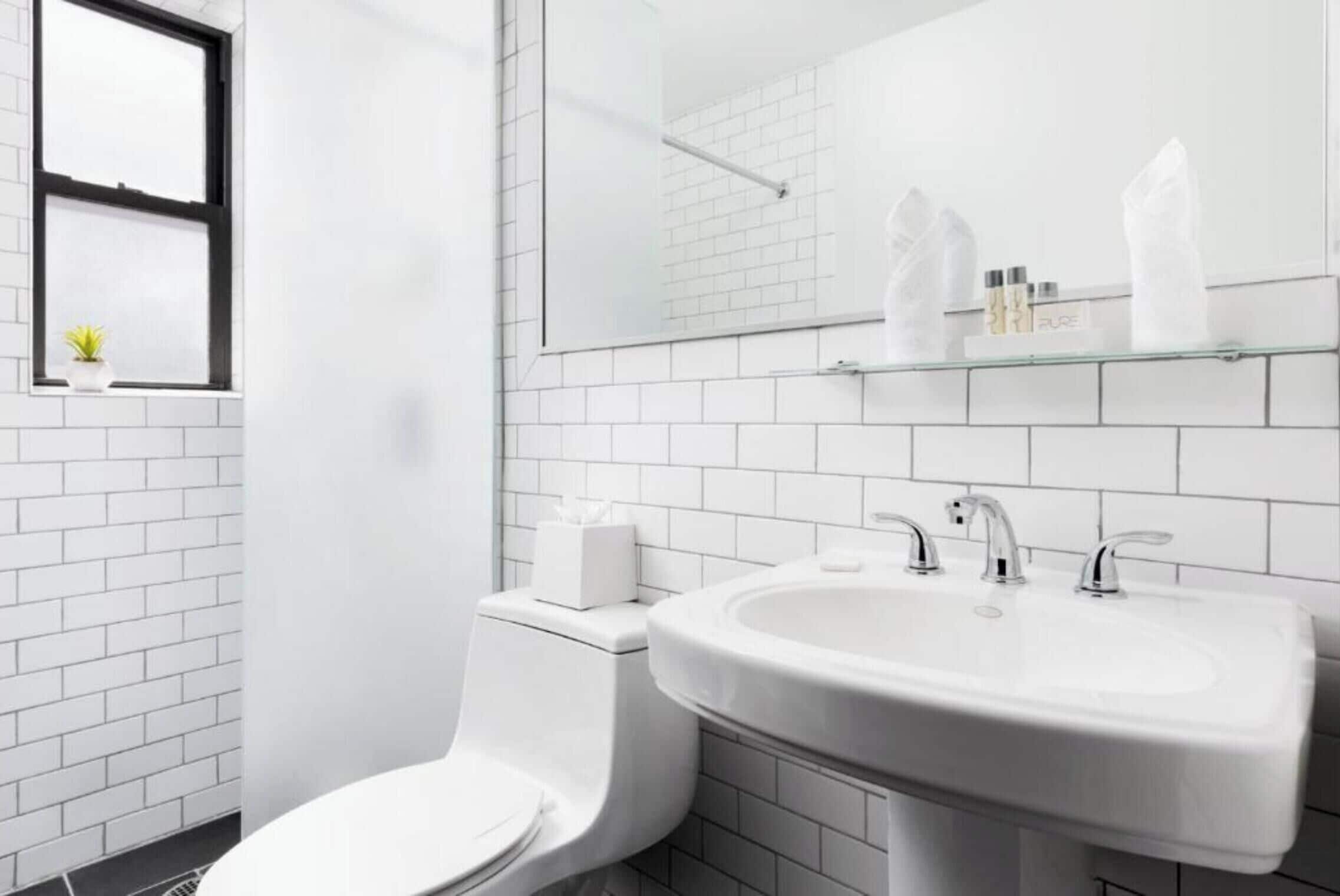 A modern bathroom with white tiles, a toilet, a sink, and a window with a plant.