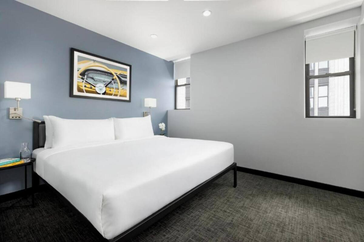 A hotel room with a large bed, two bedside tables, a framed picture on the wall, and a window with blinds.