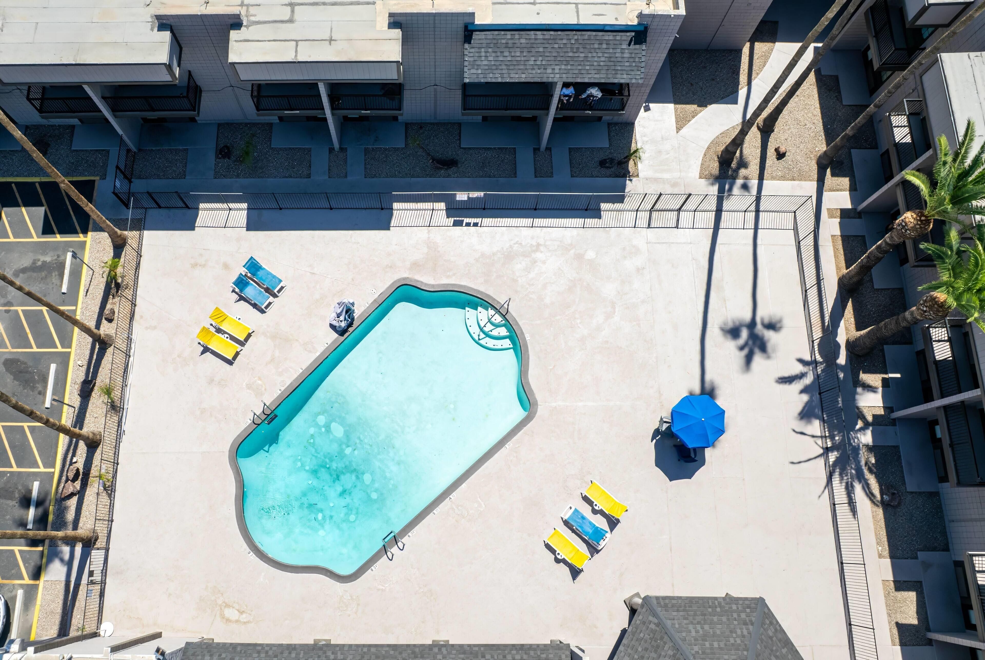 Vista aérea de uma piscina com espreguiçadeiras e um guarda-sol azul, cercada por um edifício com varandas.