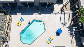 Vista aérea de uma piscina com espreguiçadeiras e um guarda-sol azul, cercada por um edifício com varandas.