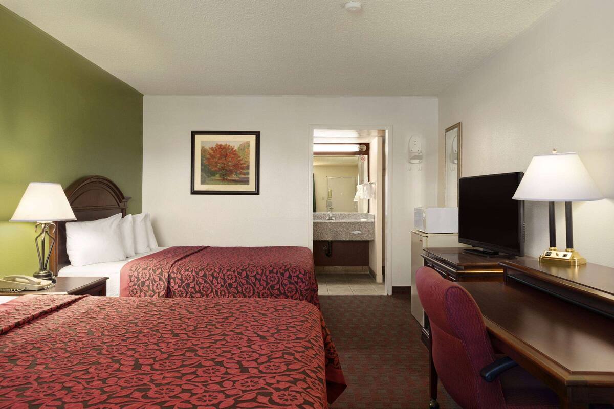 Habitación de hotel con dos camas, un escritorio con televisor, una silla, una lámpara y un baño visible a través de una puerta abierta.