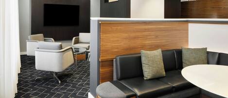 A modern hotel lobby with a black and white patterned carpet, a black leather sofa with cushions, a white round table, and a flat-screen TV mounted on the wall.