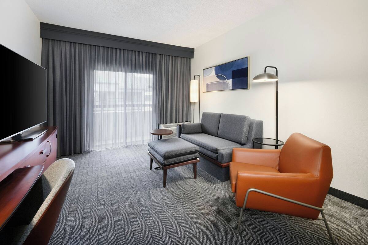 A hotel room with a sofa, ottoman, orange chair, TV, lamp, and a large window with curtains.