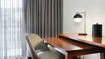 Une chambre d’hôtel avec un bureau en bois, une chaise, une lampe et une fenêtre ornée de rideaux légers.