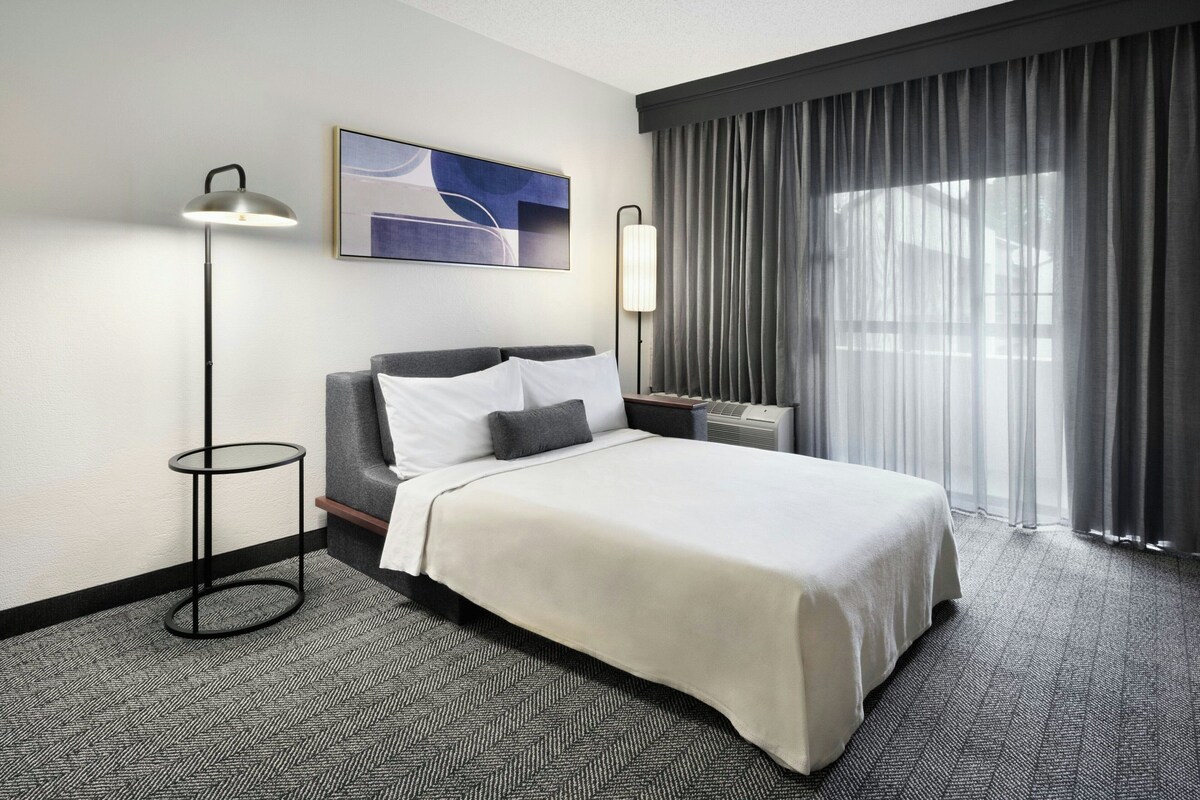 A hotel room with a large bed, a standing lamp, a painting on the wall, and a window with curtains.