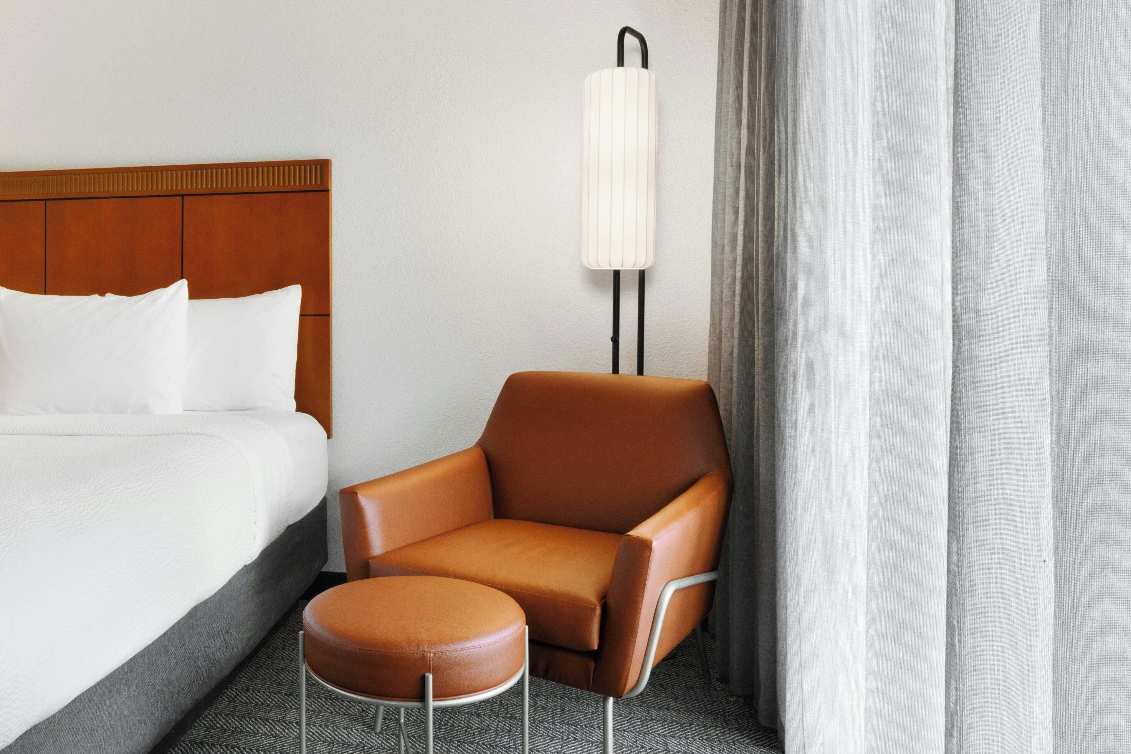 A hotel room with a bed, a brown leather chair, a small ottoman, and a wall-mounted lamp.