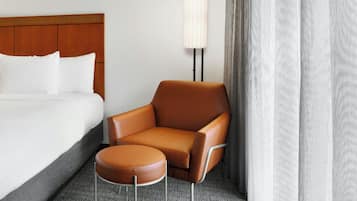 A hotel room with a bed, a brown leather chair, a small ottoman, and a wall-mounted lamp.