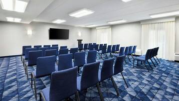 A conference room with rows of blue chairs, a flat-screen TV on the wall, and a patterned carpet.