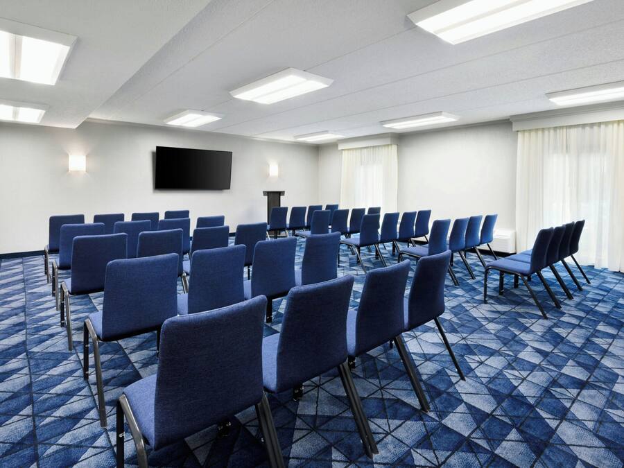 A conference room with rows of blue chairs, a flat-screen TV on the wall, and a patterned carpet.