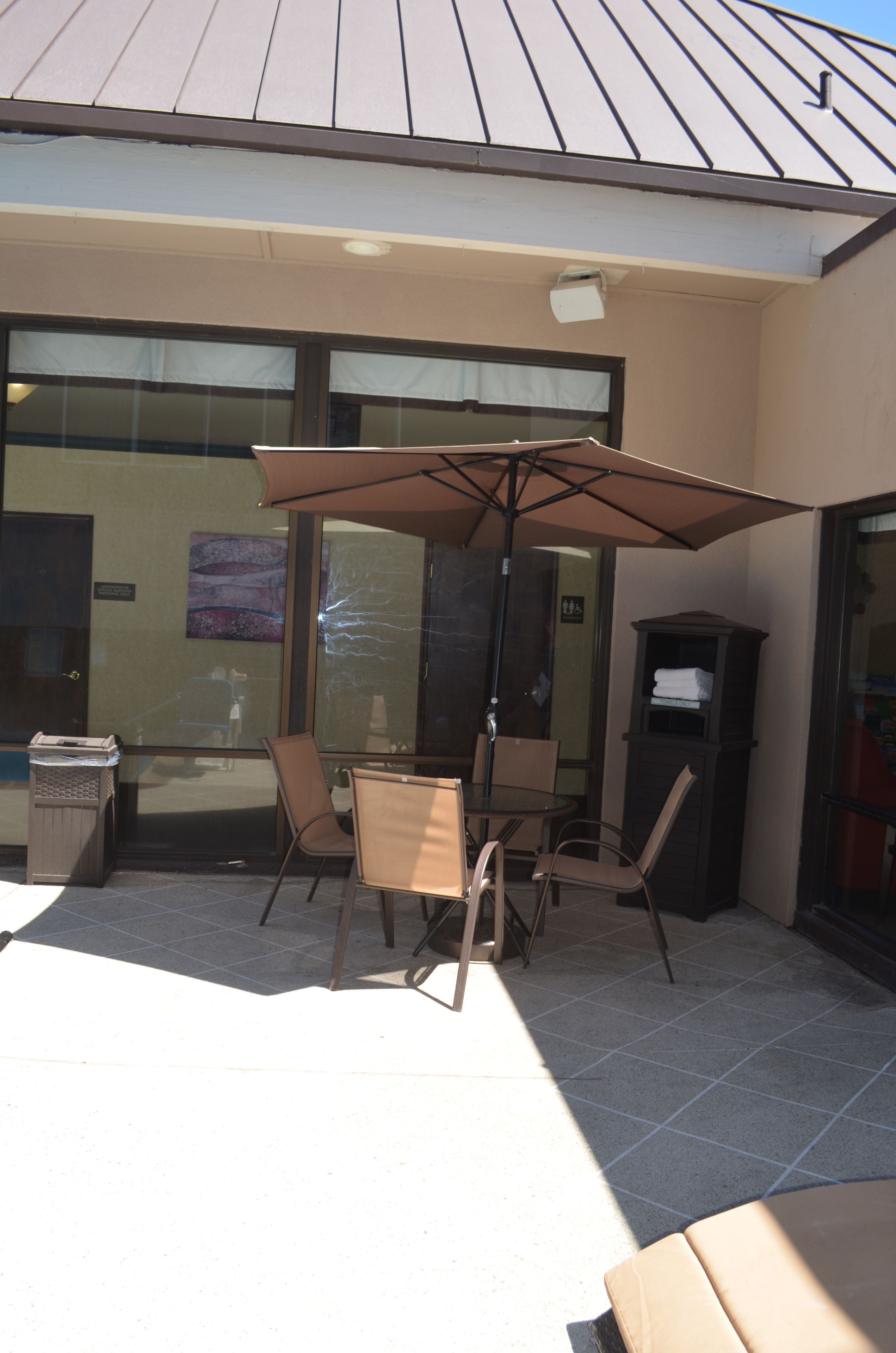 An outdoor seating area with a table and chairs under a large umbrella, a trash bin, and a door to the building.