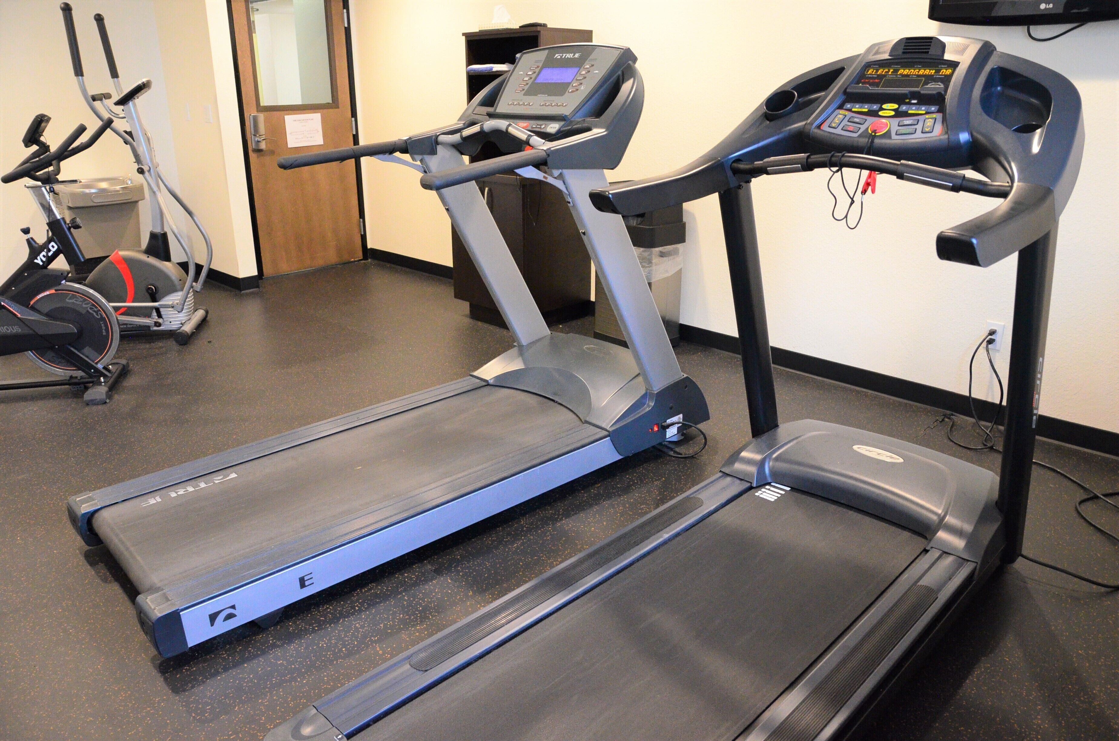 A treadmill with a digital display and control panel, located in a room with a door and a trash bin in the background.