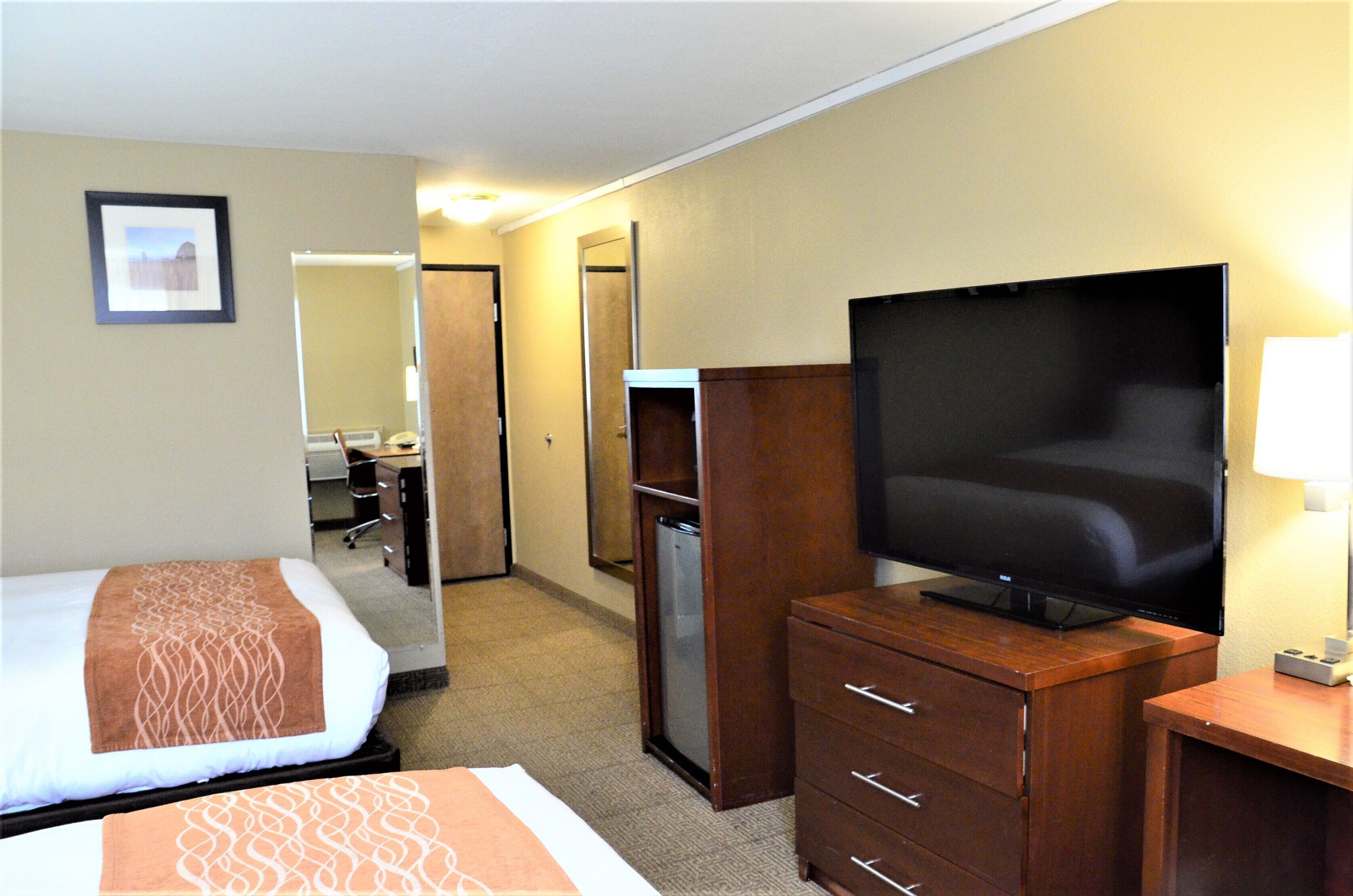 A hotel room with a bed, a television on a wooden dresser, a refrigerator, and a desk with a lamp.