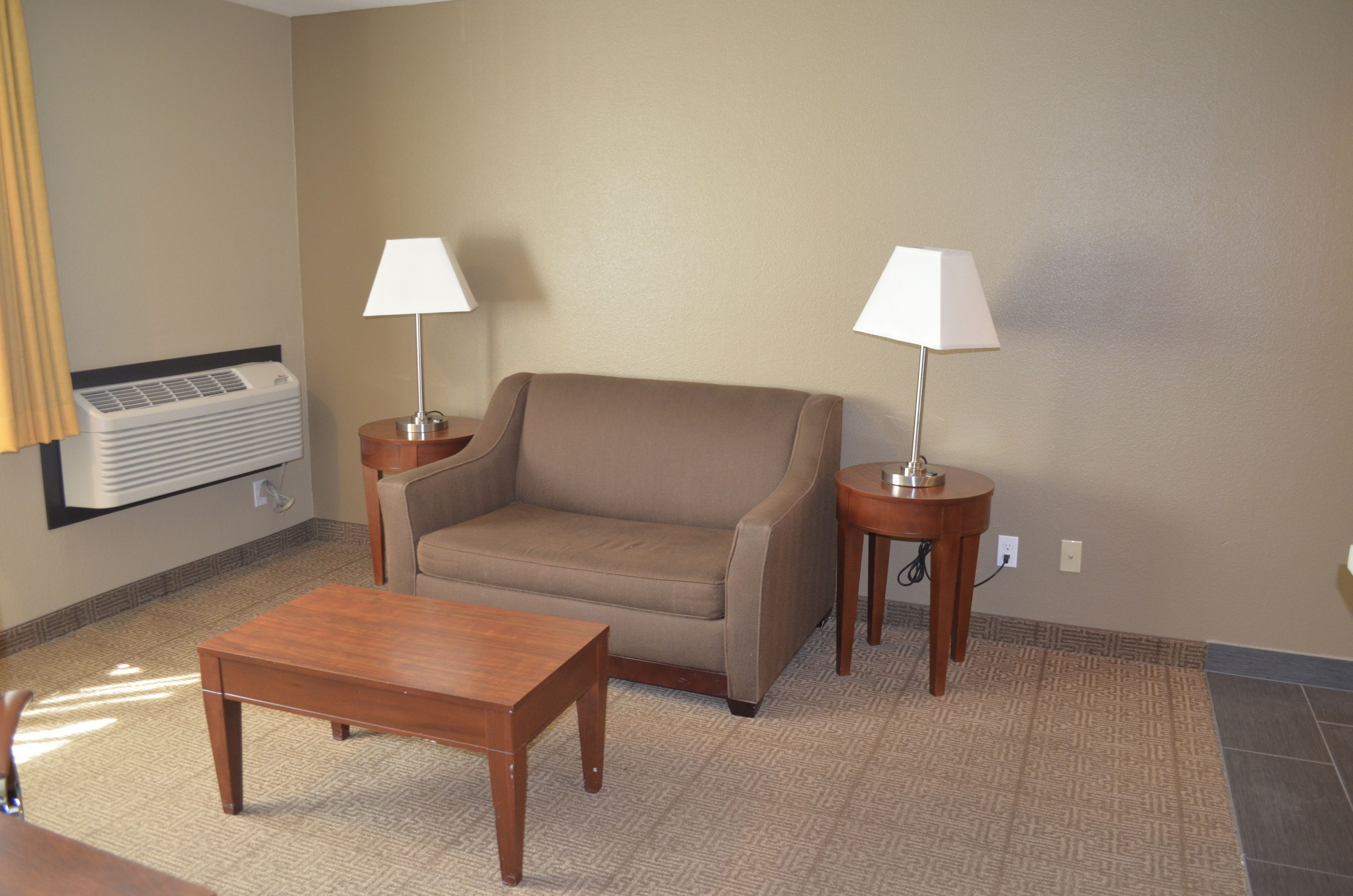 Een hotelkamer met een beige bank, twee nachtkastjes met lampen, een houten salontafel en een airconditioningunit aan de muur.