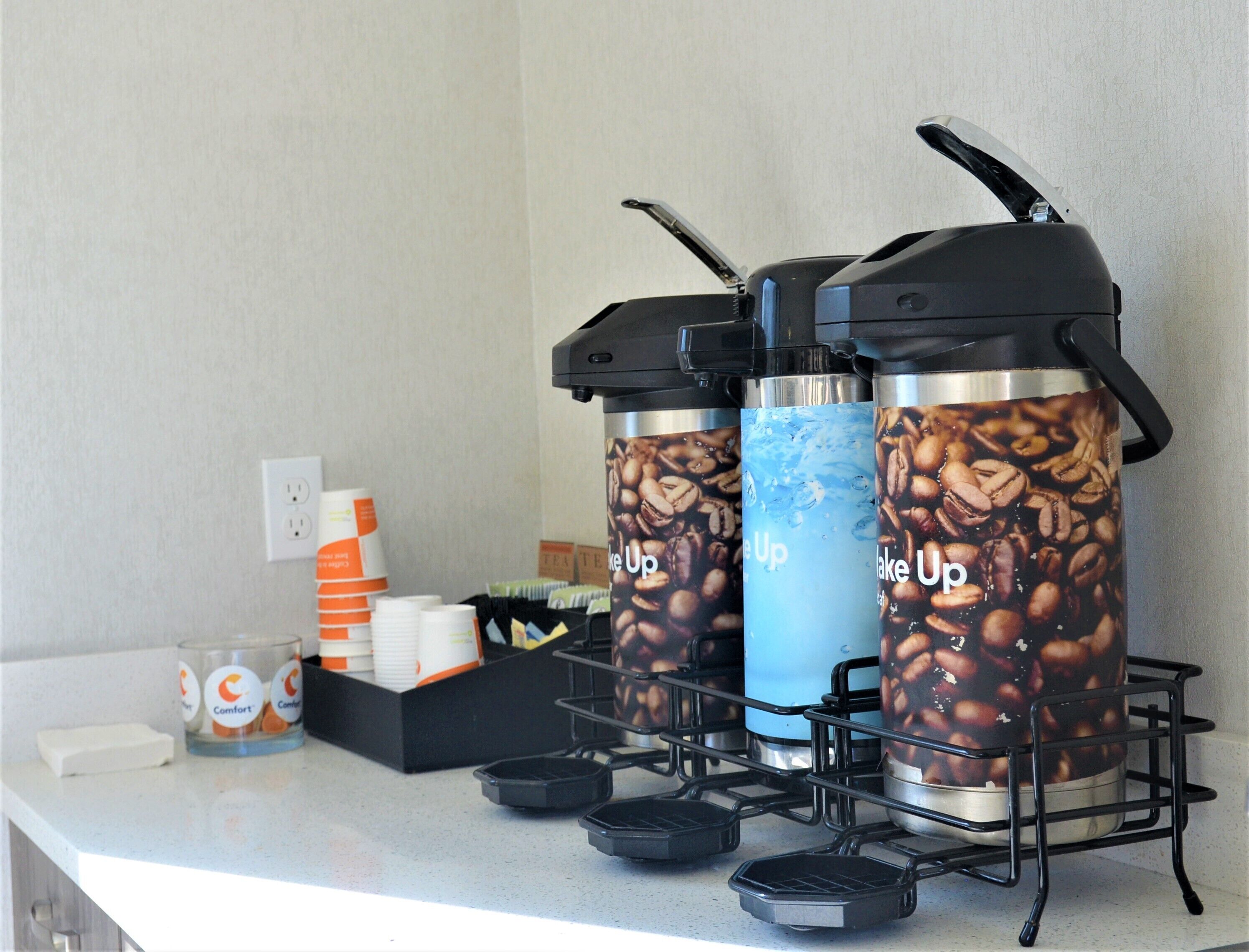 A coffee station with two coffee dispensers, a stack of disposable cups, and a small container with a coffee mug.