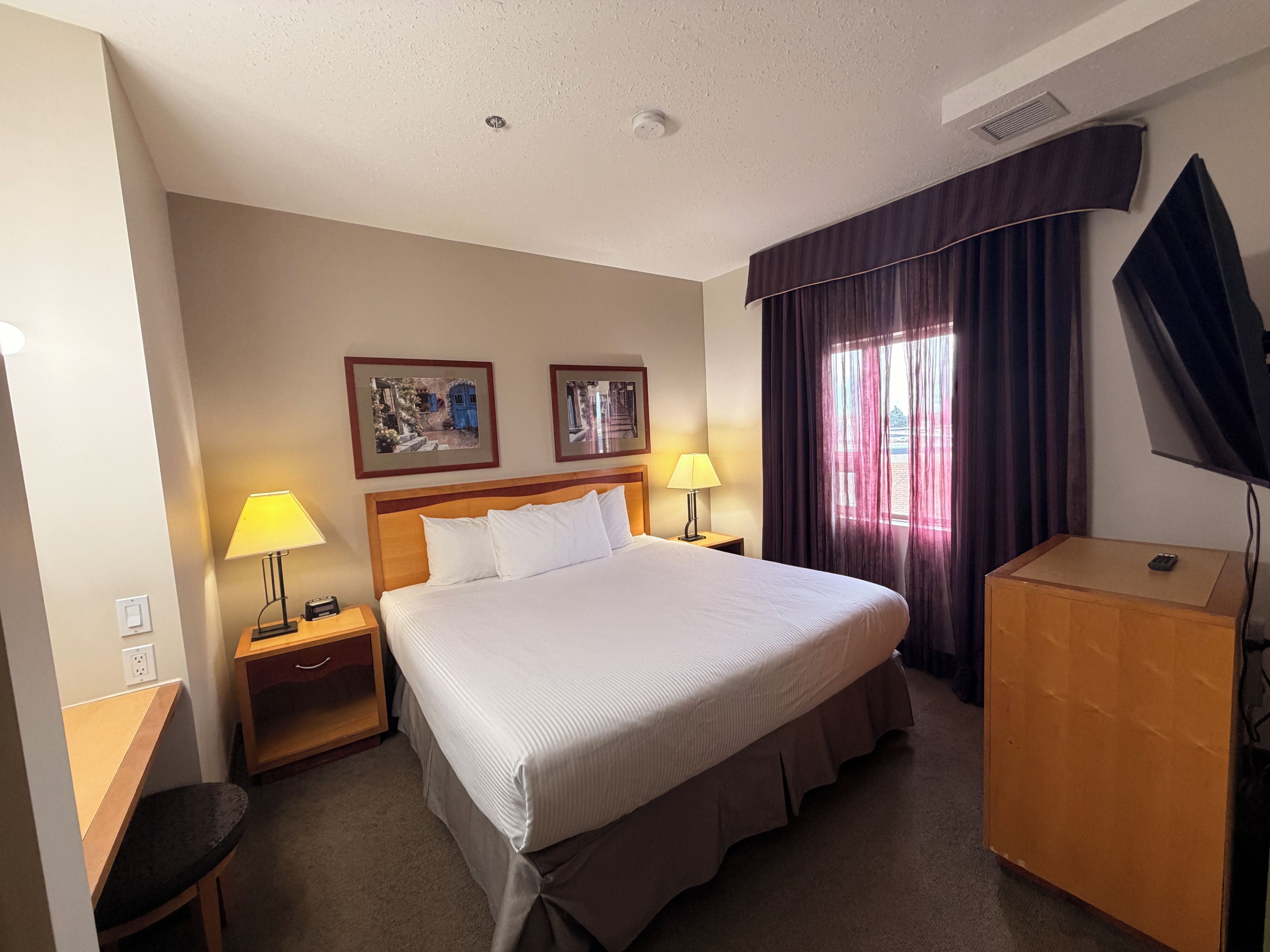 Quarto de hotel com uma cama, duas luminárias de cabeceira, uma escrivaninha, uma TV e duas fotos emolduradas na parede.