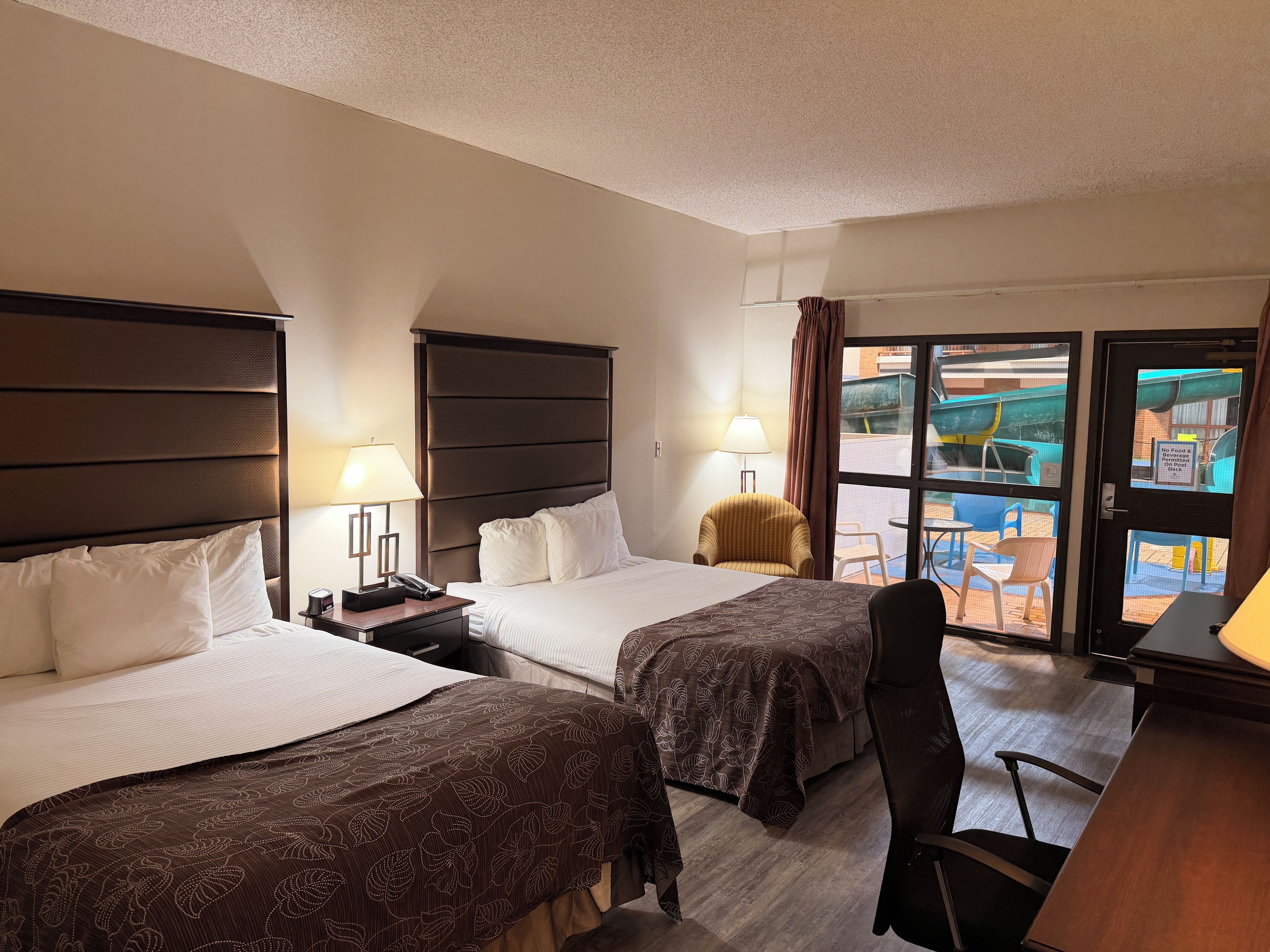 A hotel room with two beds, a desk, a chair, and a view of an outdoor area with a pool and chairs.