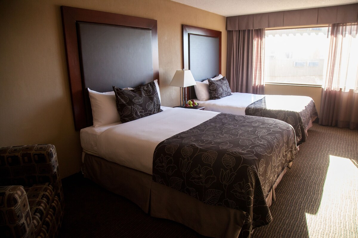 A hotel room with two beds, a nightstand with a lamp, and a view of the city through the window.
