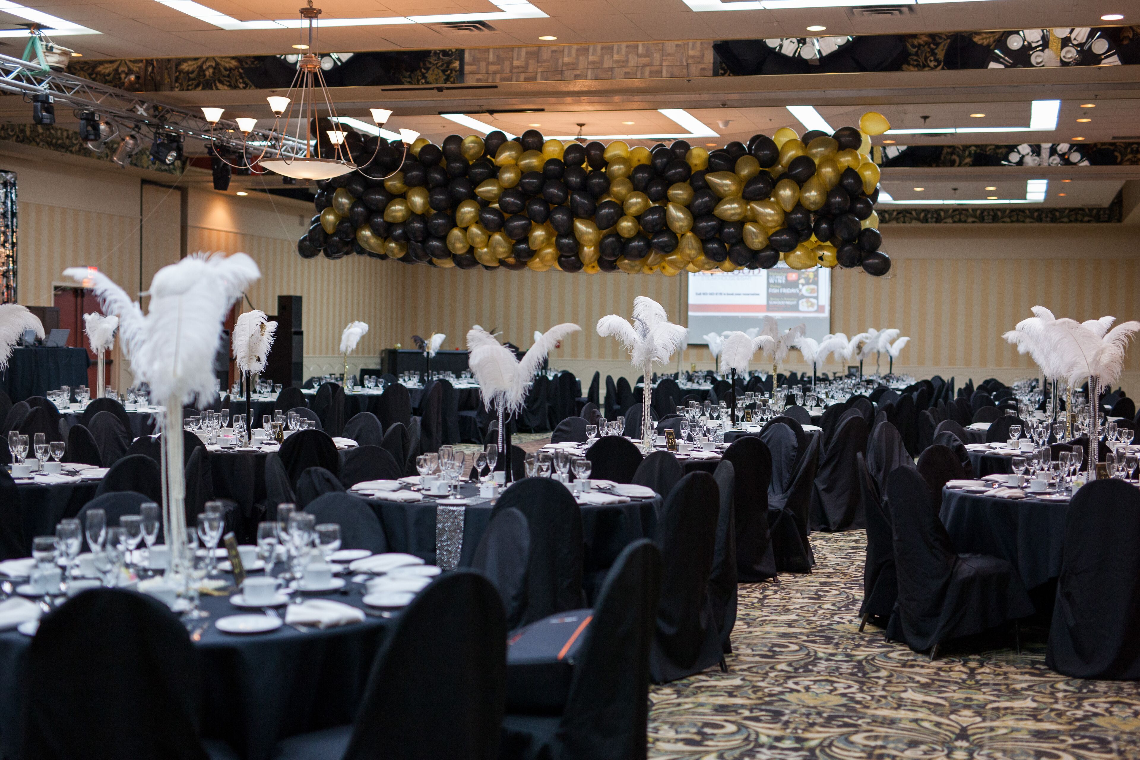 Een balzaal met ronde tafels voor een formeel evenement, versierd met zwart-wit veren en ballonnen in zwart en goud aan het plafond.