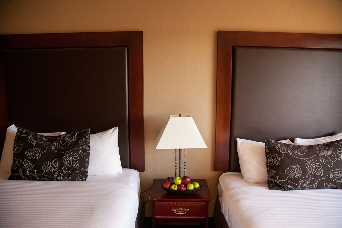 Two beds with dark headboards, a bedside table with a lamp, and a bowl of fruit.