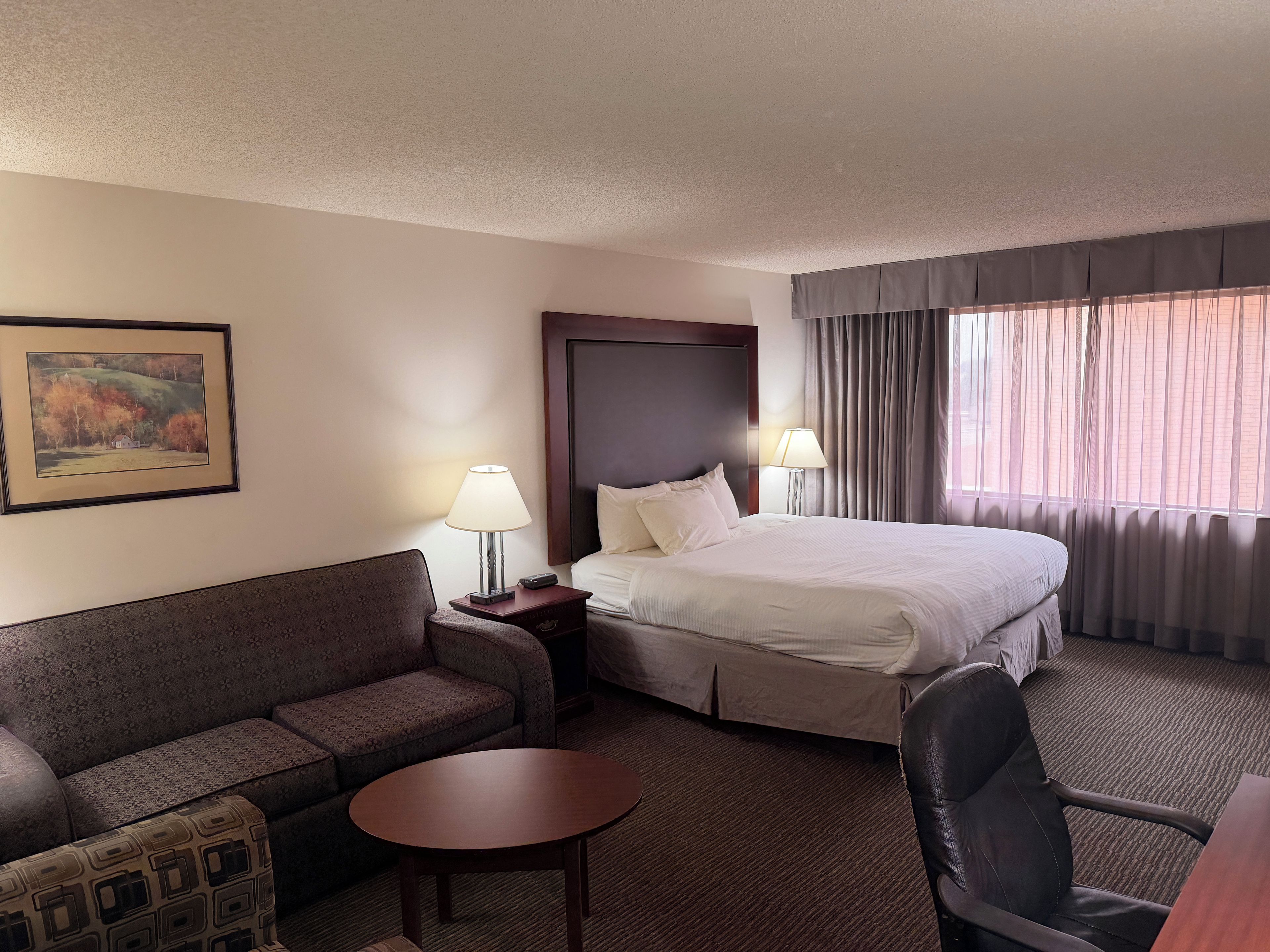 A hotel room with a bed, a sofa, a small table, a chair, and a lamp.